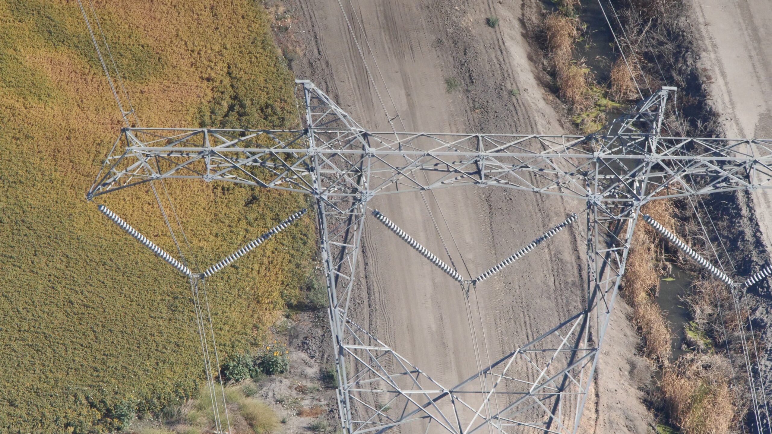 Aerial view of a tall metal power line tower over dirt paths and grassy fields, with nearby vegetation visible.
