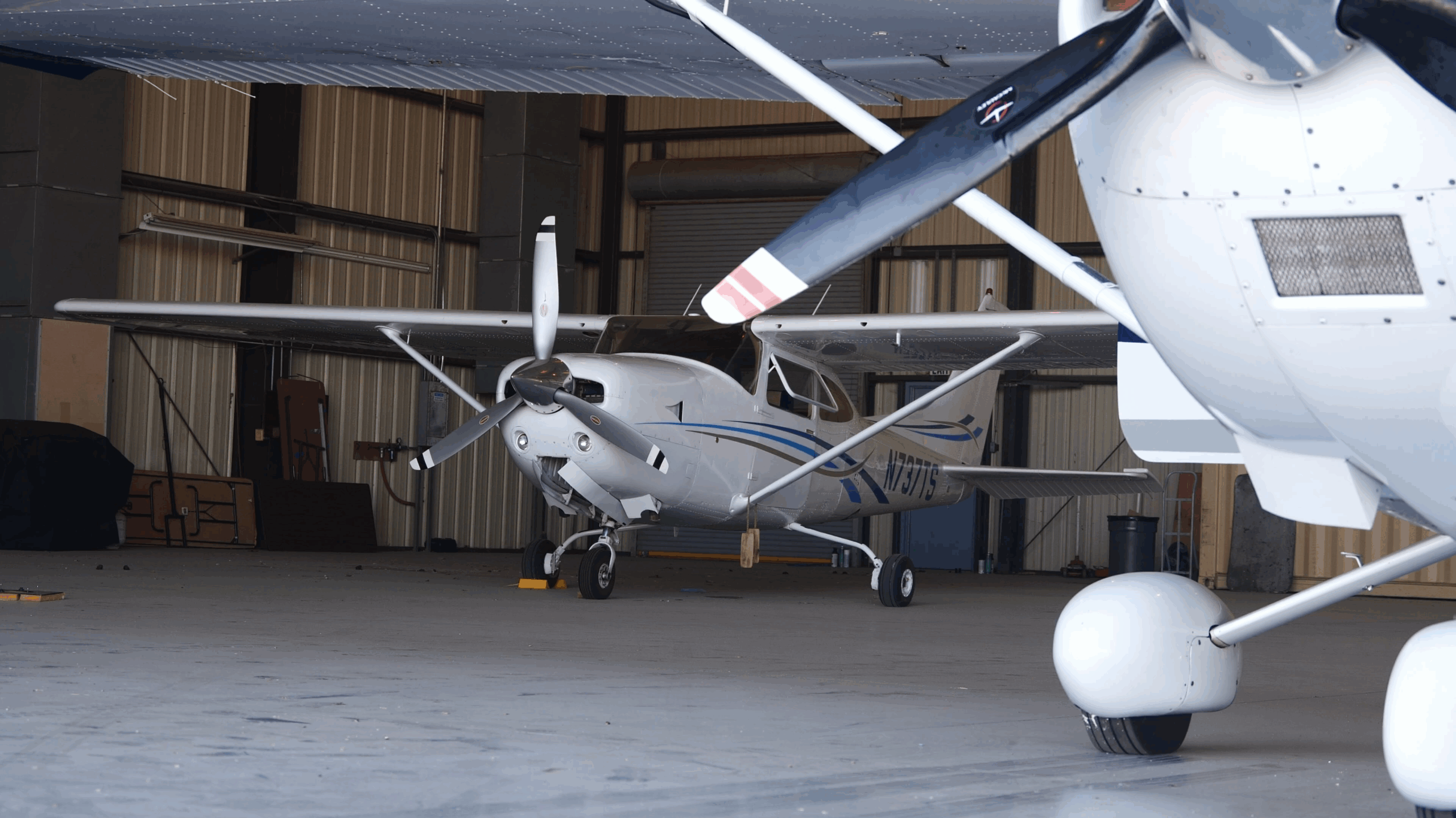 A small Cessna 182 is parked indoors in a metal hangar alongside another plane.