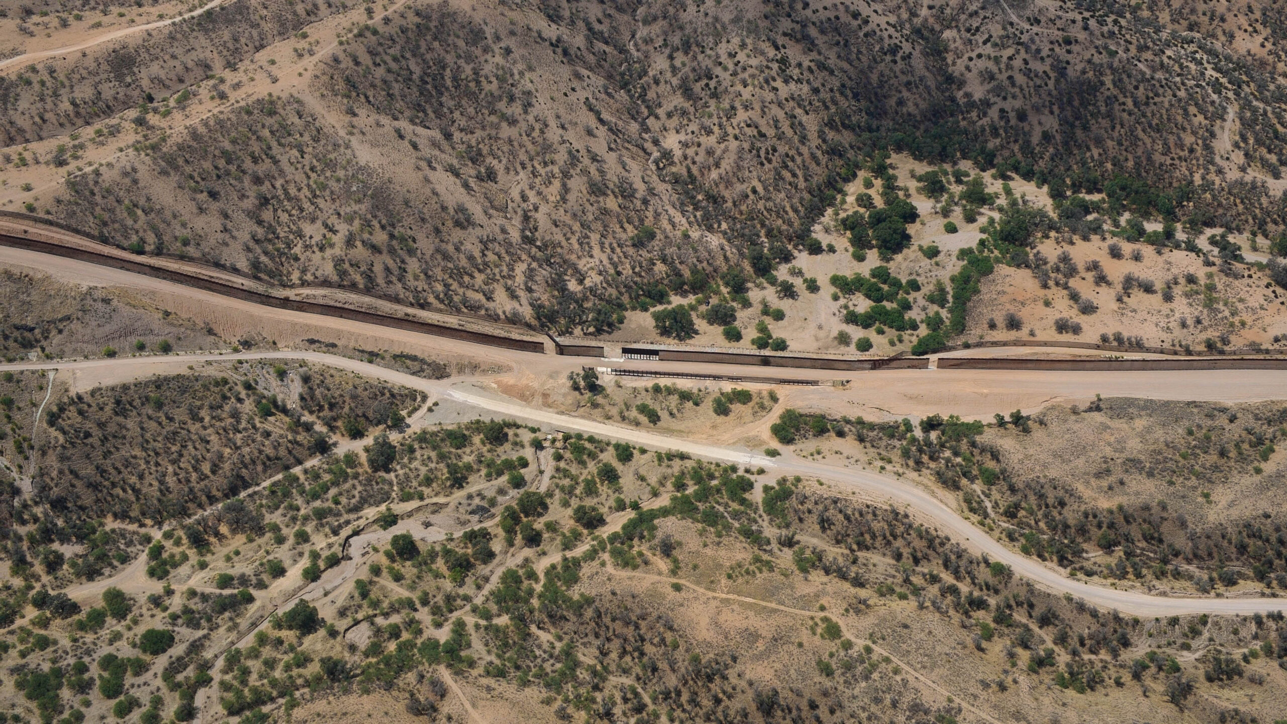 Aerial view of a desert landscape with the U.S./Mexico border wall, dirt roads, sparse vegetation, and a vehicle near the wall.