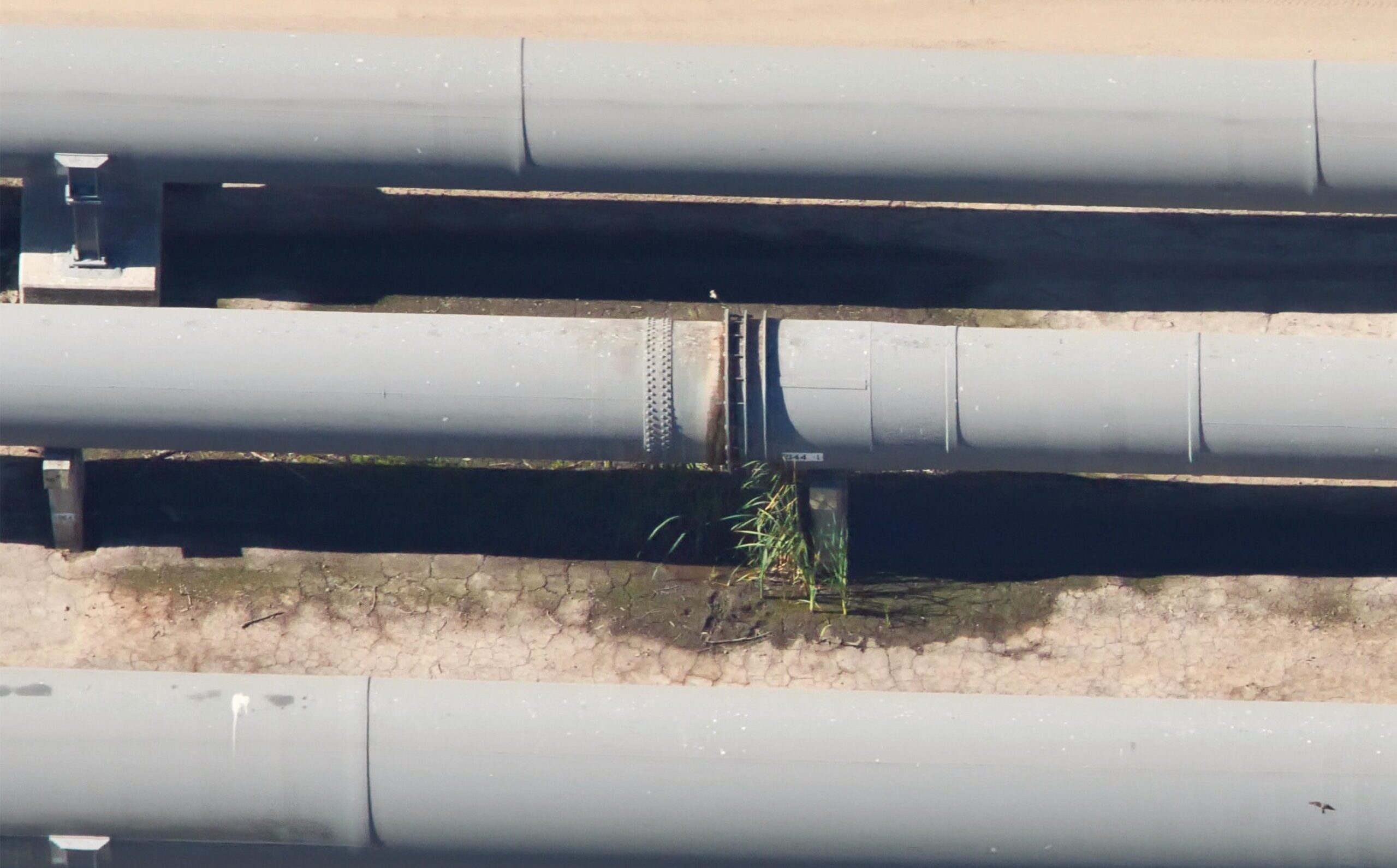 Aerial view of large gray pipelines with a cracked surface and sparse greenery in between, set against a dirt road backdrop.