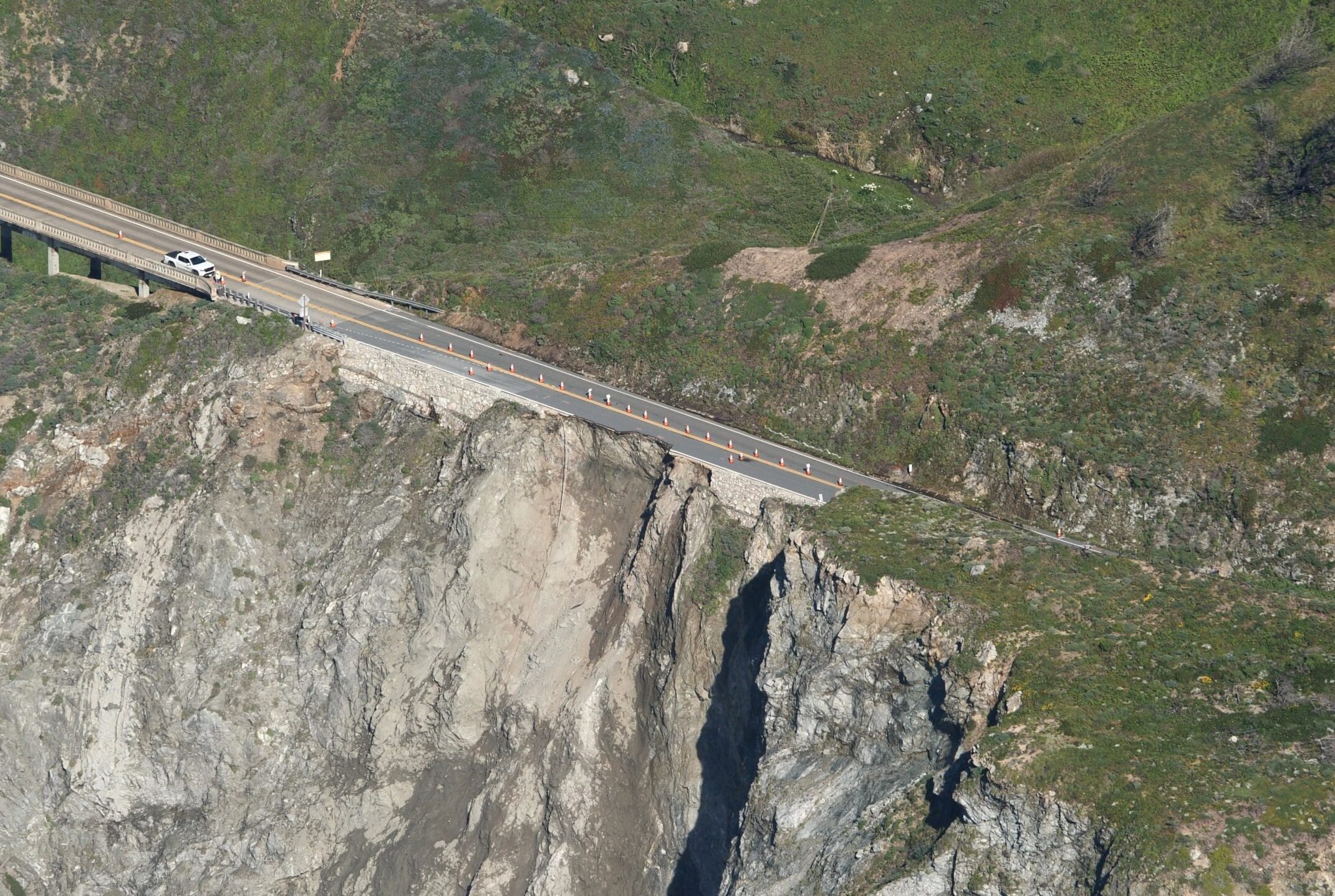 Aerial view of a highway along a steep cliff, with a vehicle and traffic cones marking construction or repair work on the edge.